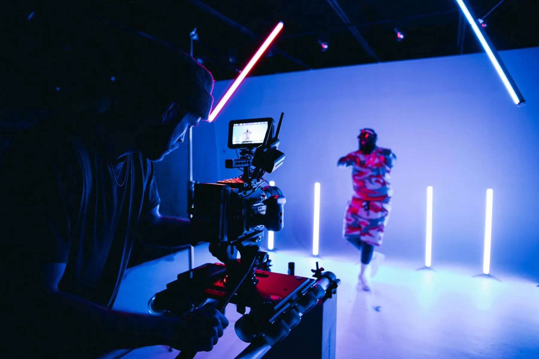 A camera operator filming a dancer on a studio set lit with neon tubes